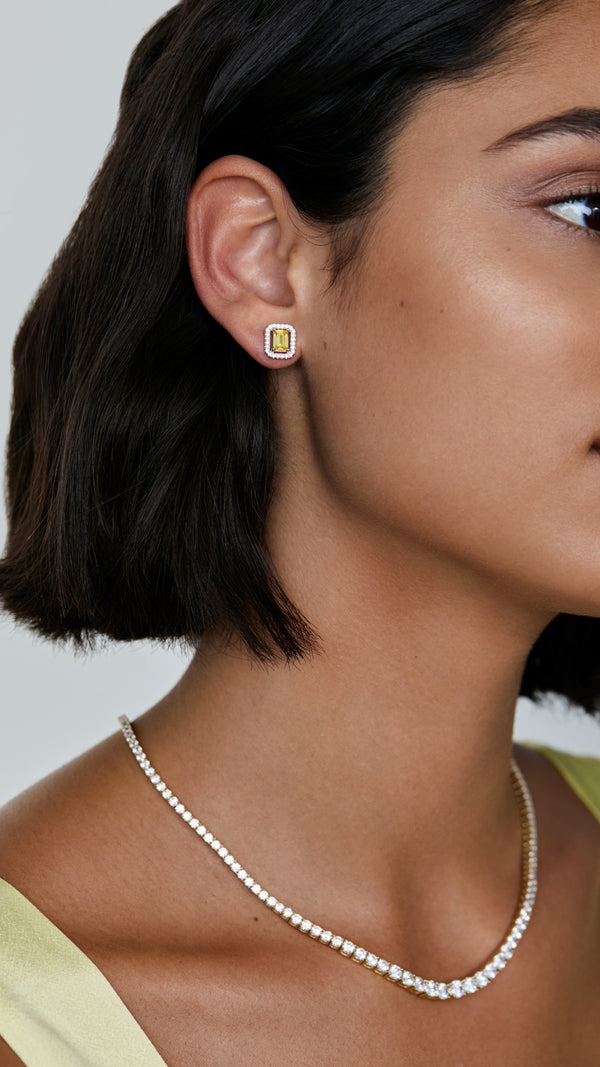 Close-up of a woman wearing canary yellow emerald cut stud earrings and a silver tennis necklace.