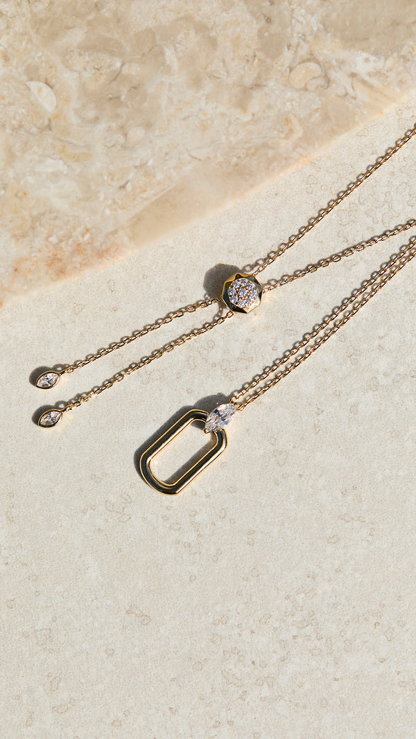 Gold necklaces with a marquise cut diamond style gemstone on a beige stone surface.