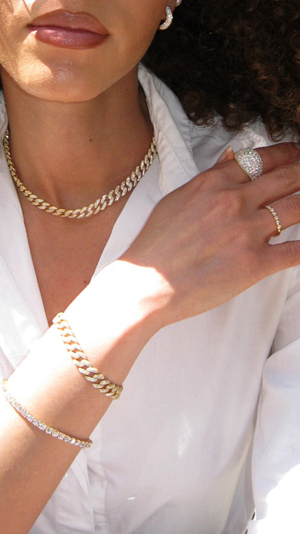 Close-up of a person wearing gold jewelry including a cuban chain necklace, bracelet, tennis bracelet and chunky pavé ring.