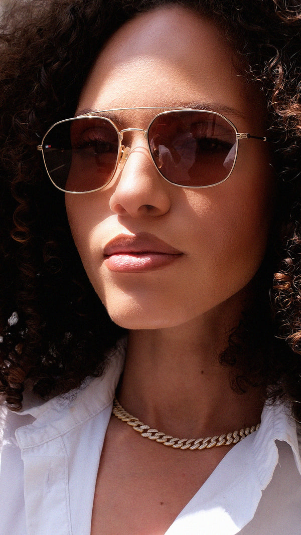 Woman wearing gold sunglasses and a white shirt with a gold cuban chain necklace.