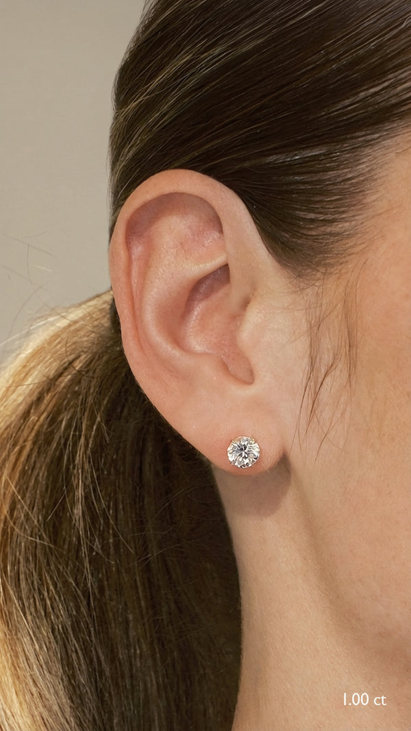 Model wearing classic 1.00 carat round brilliant solitaire earrings in 9K yellow gold, highlighting the timeless tapered basket setting and flush look on the ear.