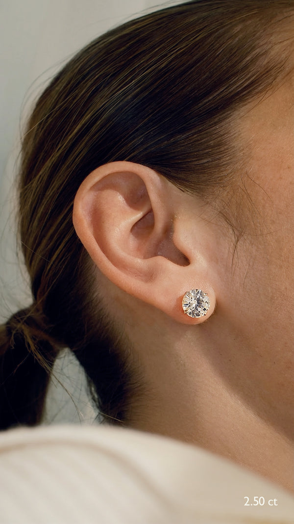 A close-up of a model wearing everyday 2.5 carat round brilliant solitaire stud earrings in 9K yellow gold, demonstrating the secure, flush fit on the earlobe.