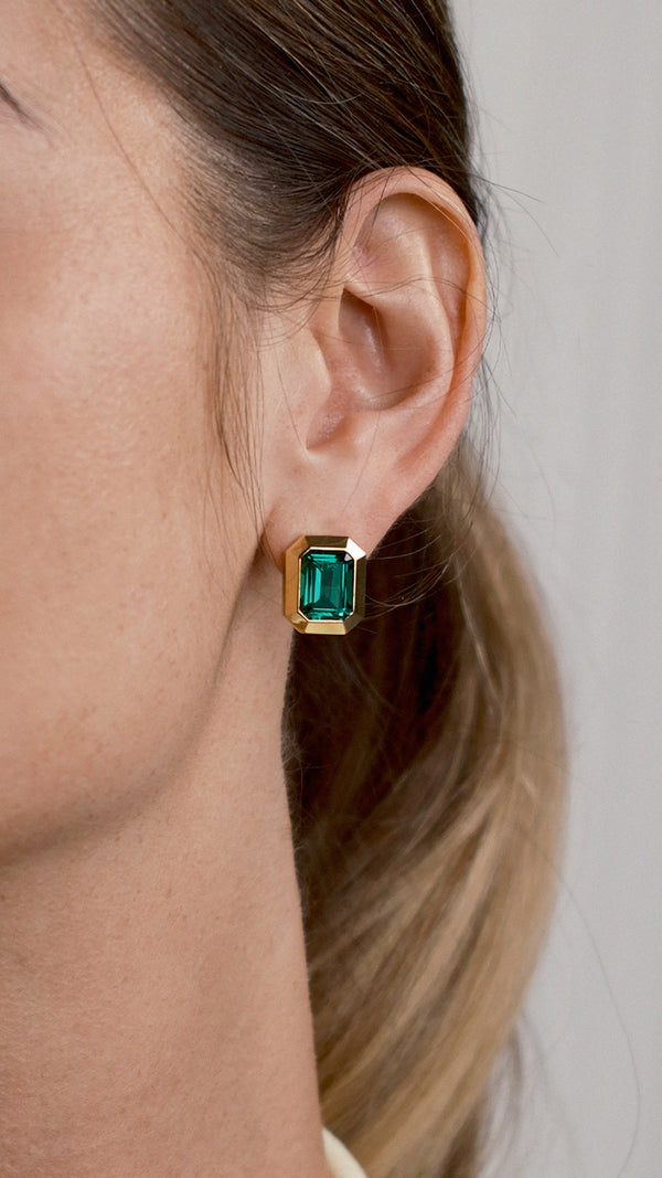 Close-up of an ear wearing a green gemstone earring with gold setting against a neutral background