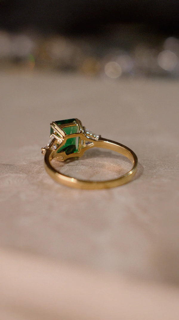 Gold ring with a green radiant cut emerald gemstone on a neutral background
