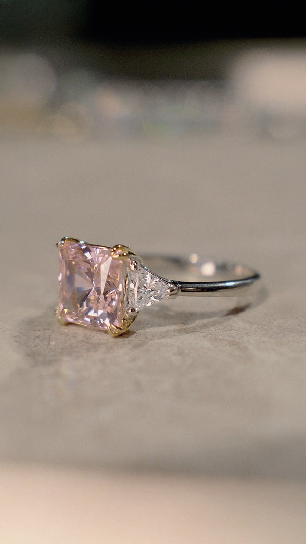 A princess-cut diamond style trilogy ring with a pink gemstone.