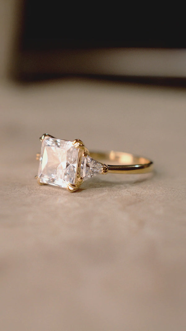 Gold ring with a large princess-cut diamond style gemstone on a beige background.