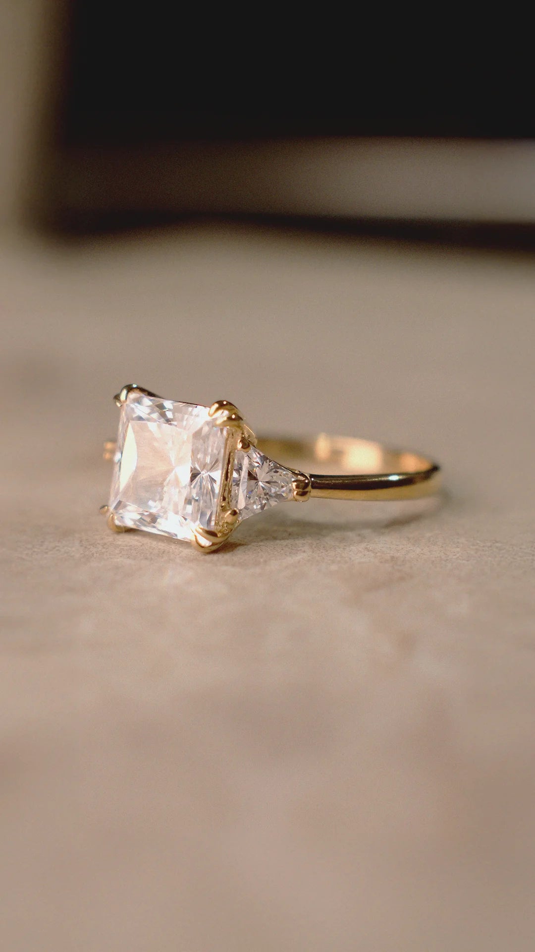 Gold ring with a large princess-cut diamond style gemstone on a beige background.