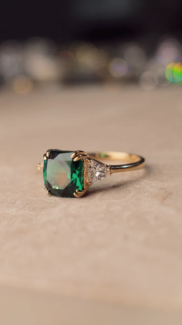 Gold ring with a green gemstone and triangular shoulder stones on a beige background.