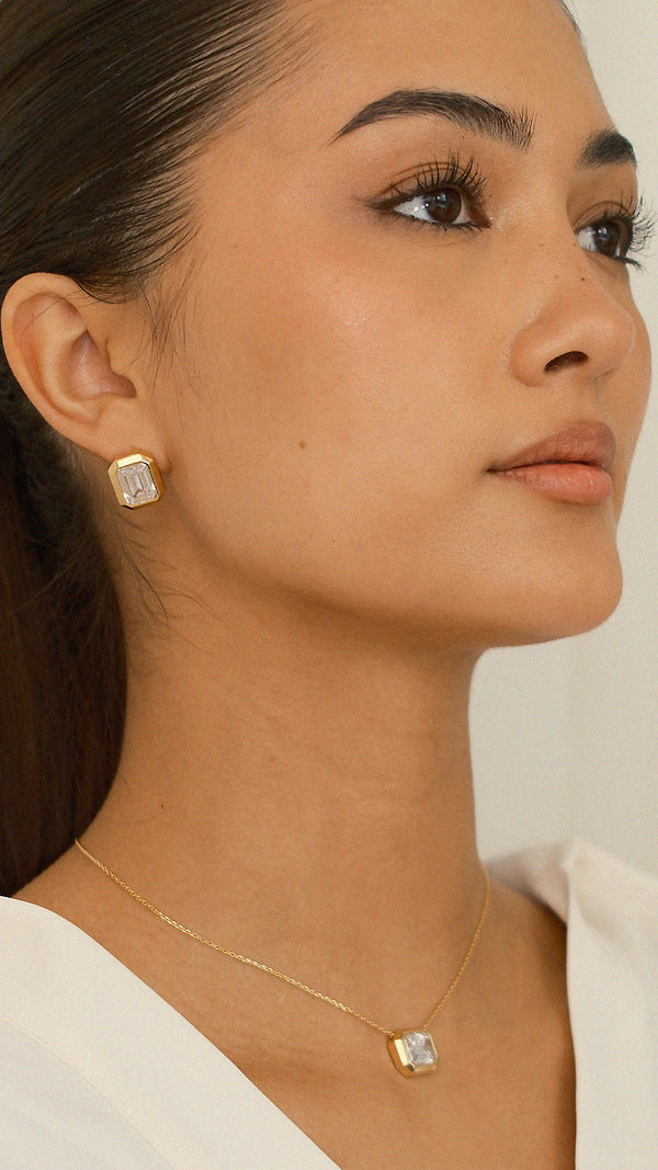 Trendy gold statement stud earrings on ear, showcasing a rectangular emerald-cut keystone with a thick gold border for high-voltage drama. Paired with matching slider necklace. 