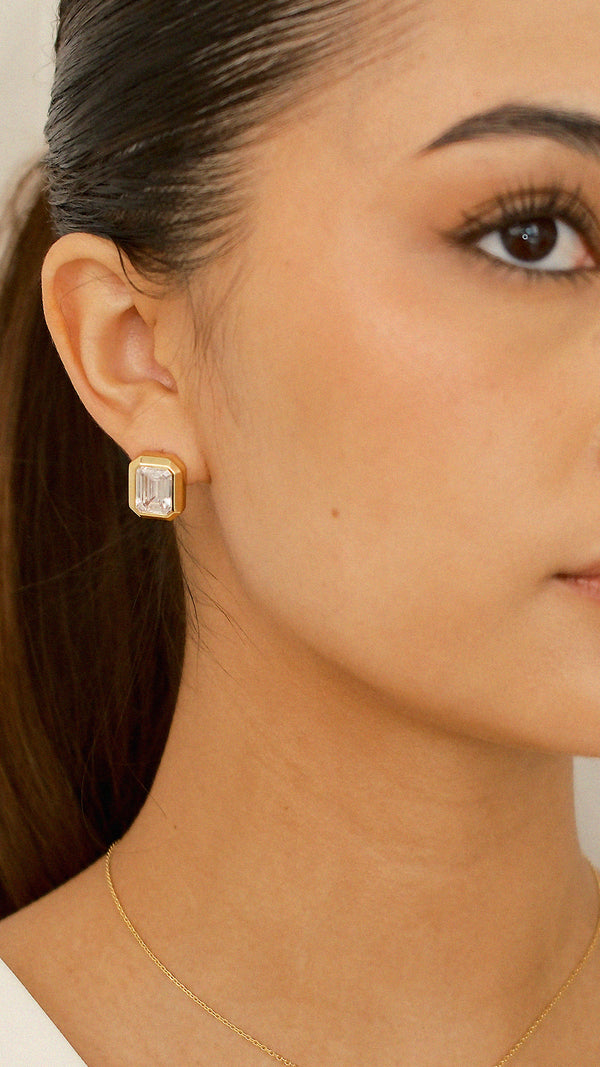 A close-up of a model wearing a gold Lily earring, featuring a large emerald-cut gemstone encased in a chunky, polished gold bezel setting.