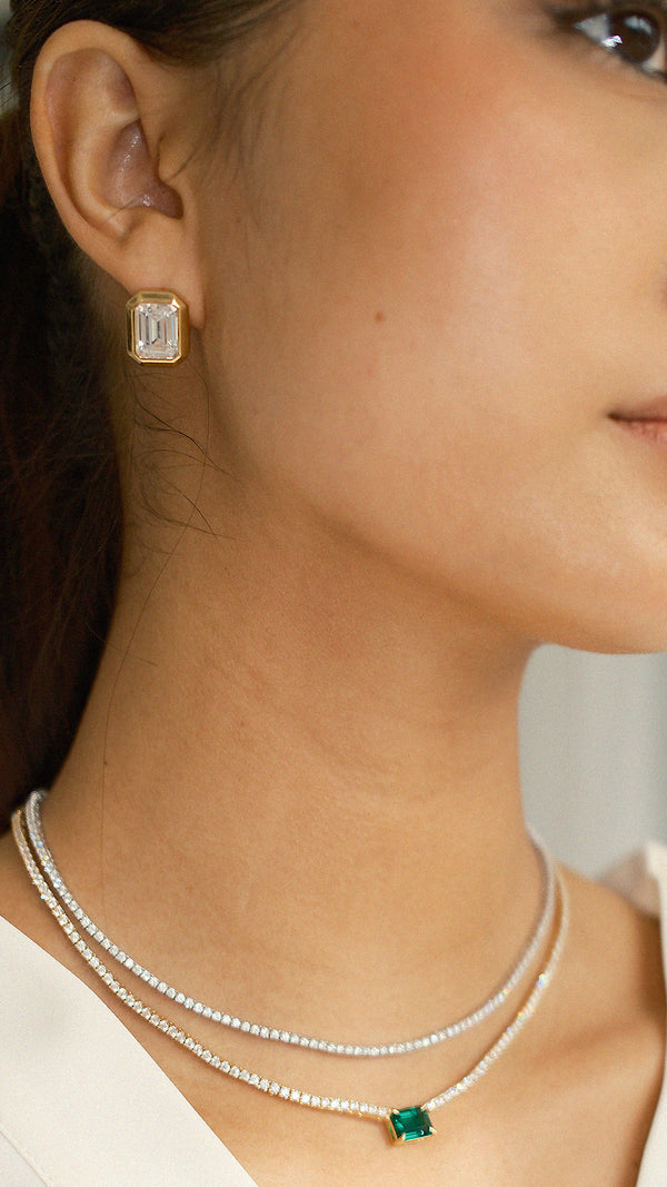 Professional shot highlighting the light-reflecting facets of a clear rectangular stud in a gold setting and a matching green emerald-cut pendant on tennis necklace chain.