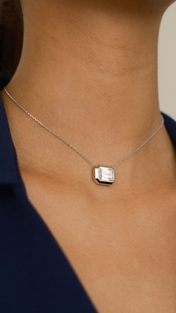 A close-up of a model wearing a silver Lily necklace featuring a clear emerald-cut gemstone in a thick, polished silver bezel setting. Styled over a navy blue collar to show length and scale.