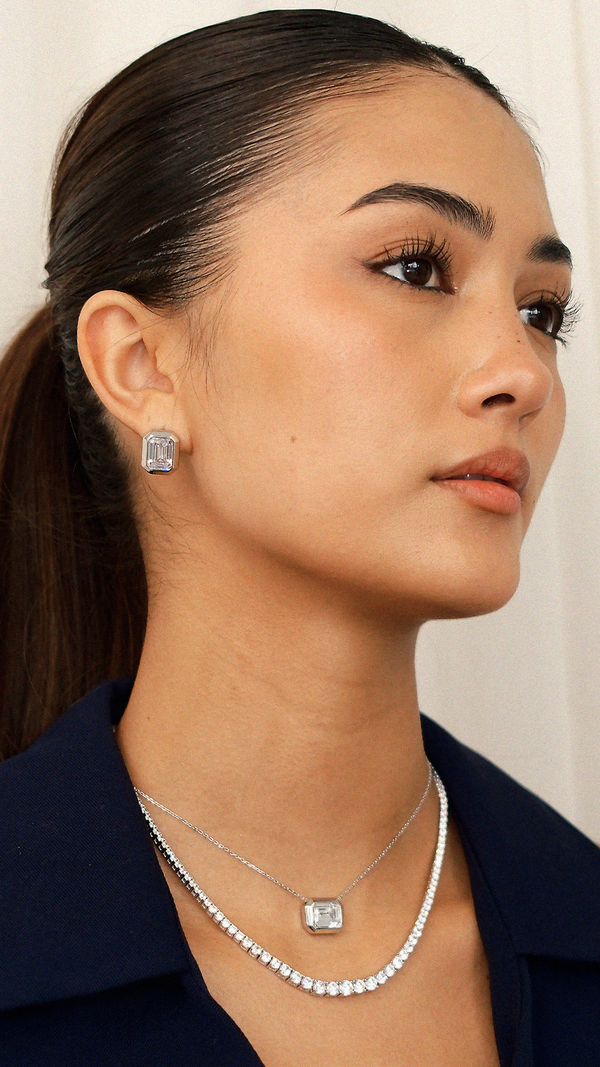 Side-profile shot of silver statement jewellery. The large, rectangular emerald-cut stud earrings are shown on the ear to illustrate scale, paired with a layered silver tennis chain and a matching bezel-set keystone necklace.