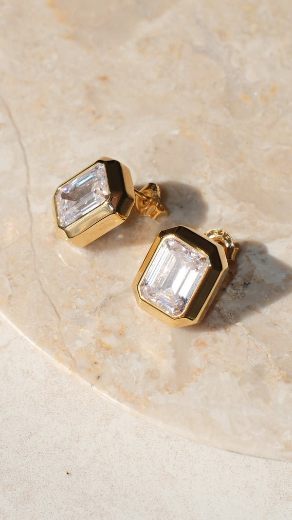 Gold earrings with clear emerald-cut gemstone on a beige stone surface