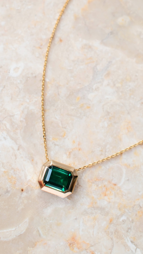Gold slider necklace with an emerald green gemstone on a marble surface