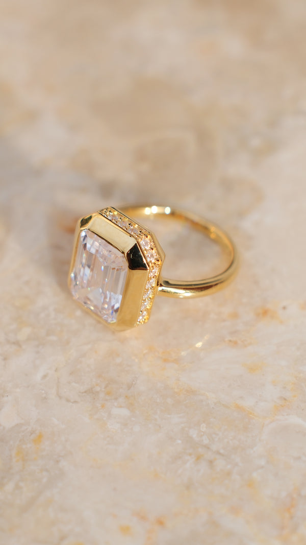 Side-profile view of a gold Lily ring on a marble surface, highlighting the large emerald-cut  gemstone in a chunky bezel setting with a hidden pavé halo of small stones underneath.