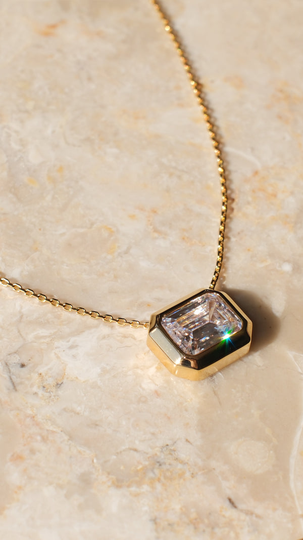 A  flat lay of the gold Lily pendant necklace on a textured marble background. The image captures the light reflecting off the emerald-cut facets and the smooth, polished finish of the chunky gold bezel.