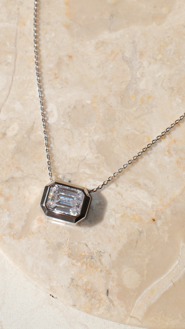 Flat lay of a silver emerald-cut pendant necklace on a marble surface, highlighting the crisp facets of the clear rectangular keystone and the durable chunky bezel.