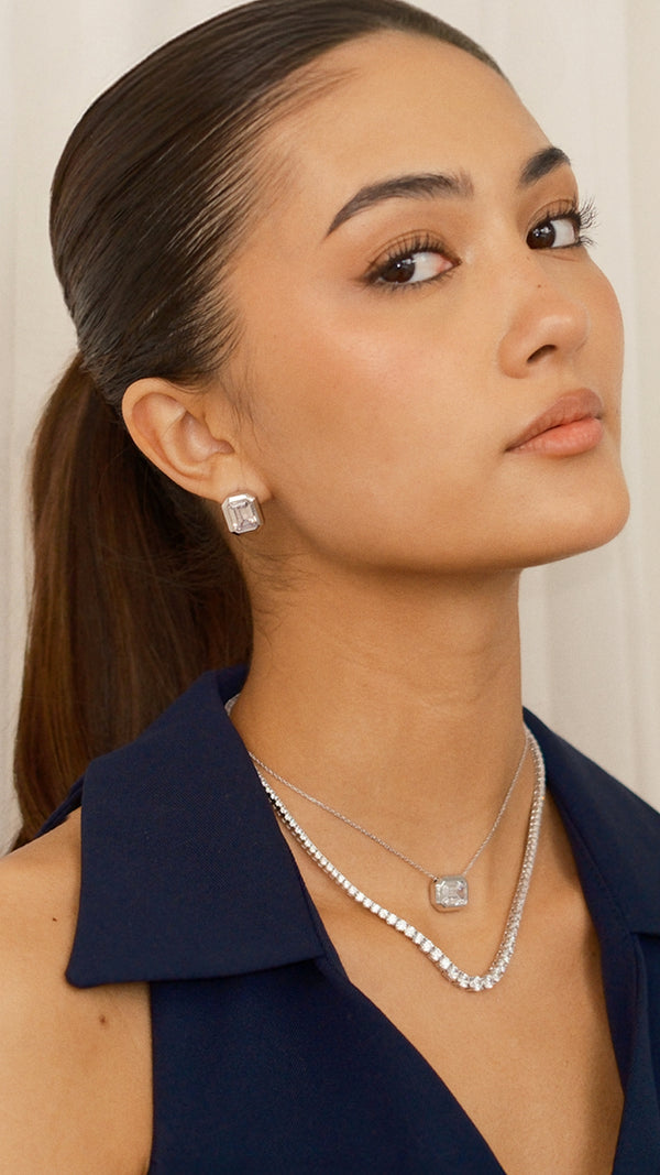 Close-up model shot of a sterling silver and clear gemstone jewellery. The model wears emerald-cut stud earrings paired with a layered silver tennis necklace and a matching bezel-set emerald-cut pendant