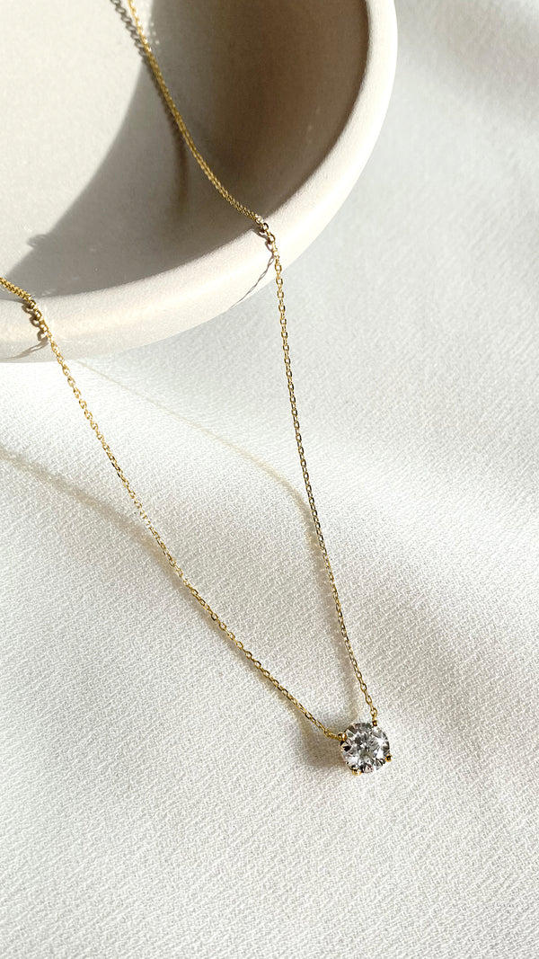 A solitaire diamond necklace set in 18K yellow gold, displayed on a white surface with a bowl in the background.