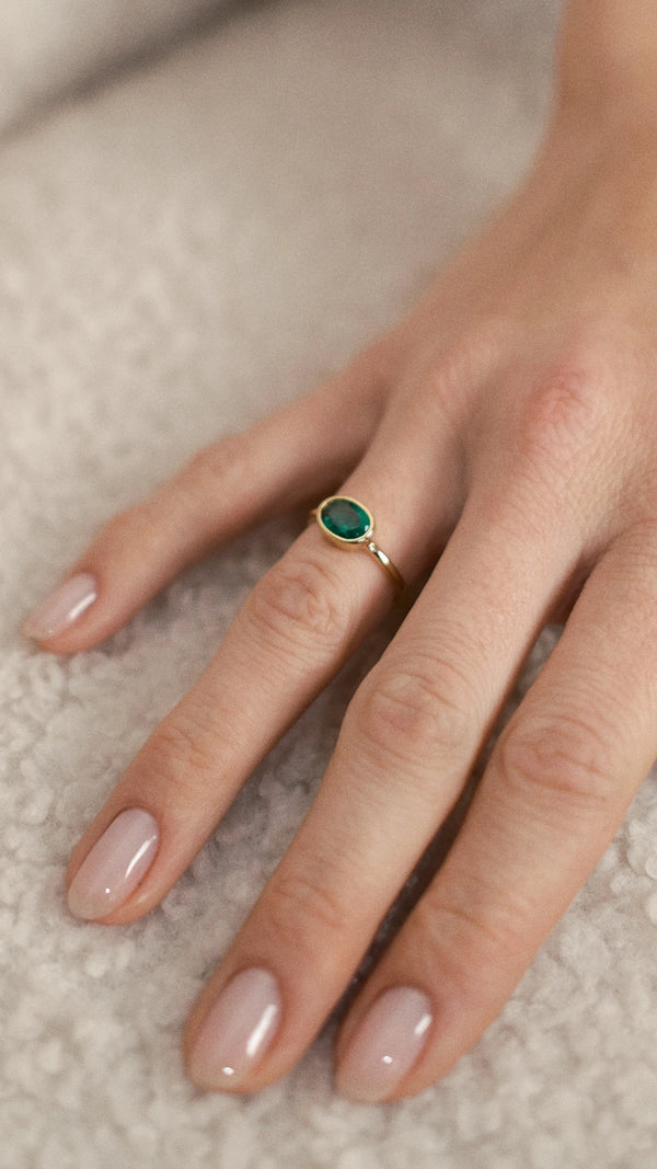 Hand wearing a gold ring with an oval green gemstone on a neutral background