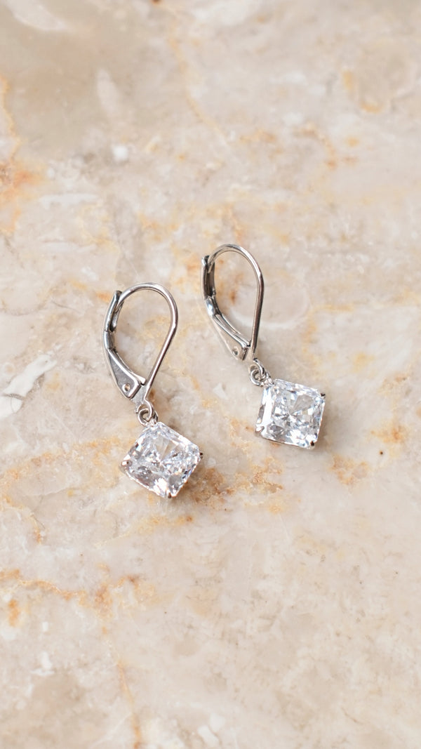 Pair of silver earrings with princess cut gemstones on a beige marble surface