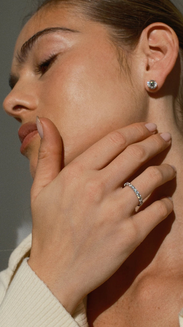 Close-up of a woman's hand with a white gold eternity ring and diamond style stud earring, wearing a beige sweater.