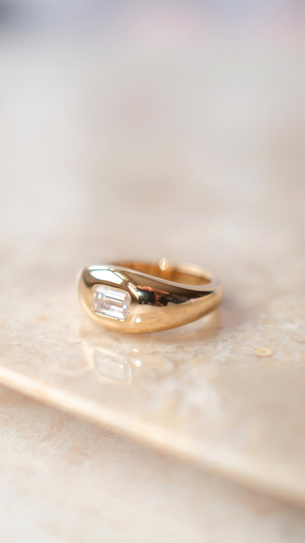 Smooth tapered gold band ring featuring a solitaire emerald cut gemstone.
