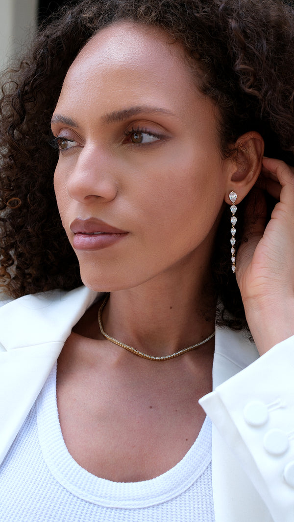Woman wearing a gold tennis necklace and drop earrings, with a close-up of her ear.