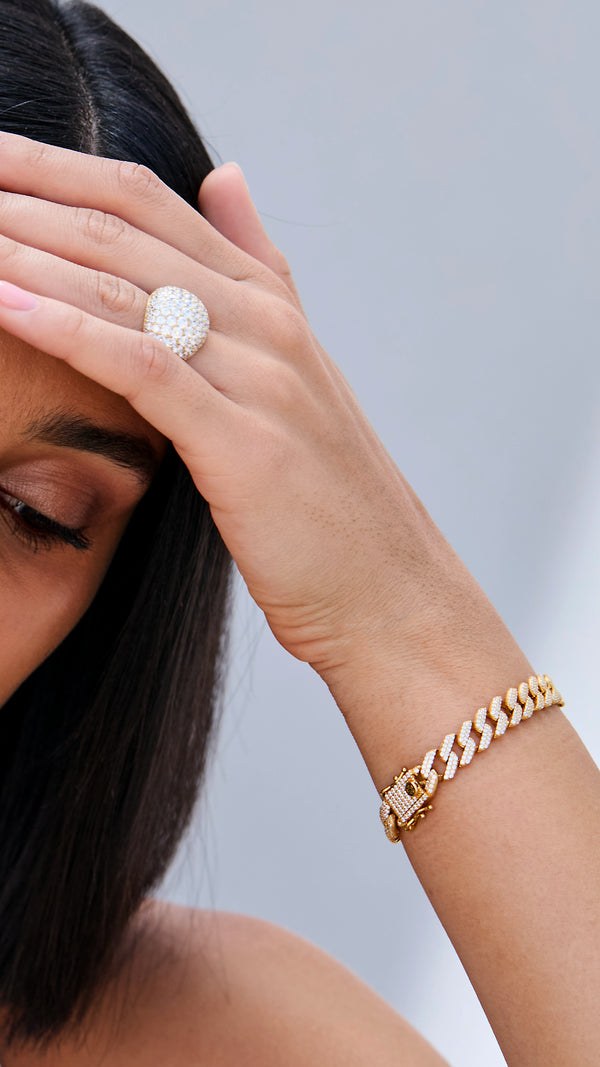 Close-up of a hand wearing a gold cuban link bracelet and ring dome shaped ring on a  neutral background