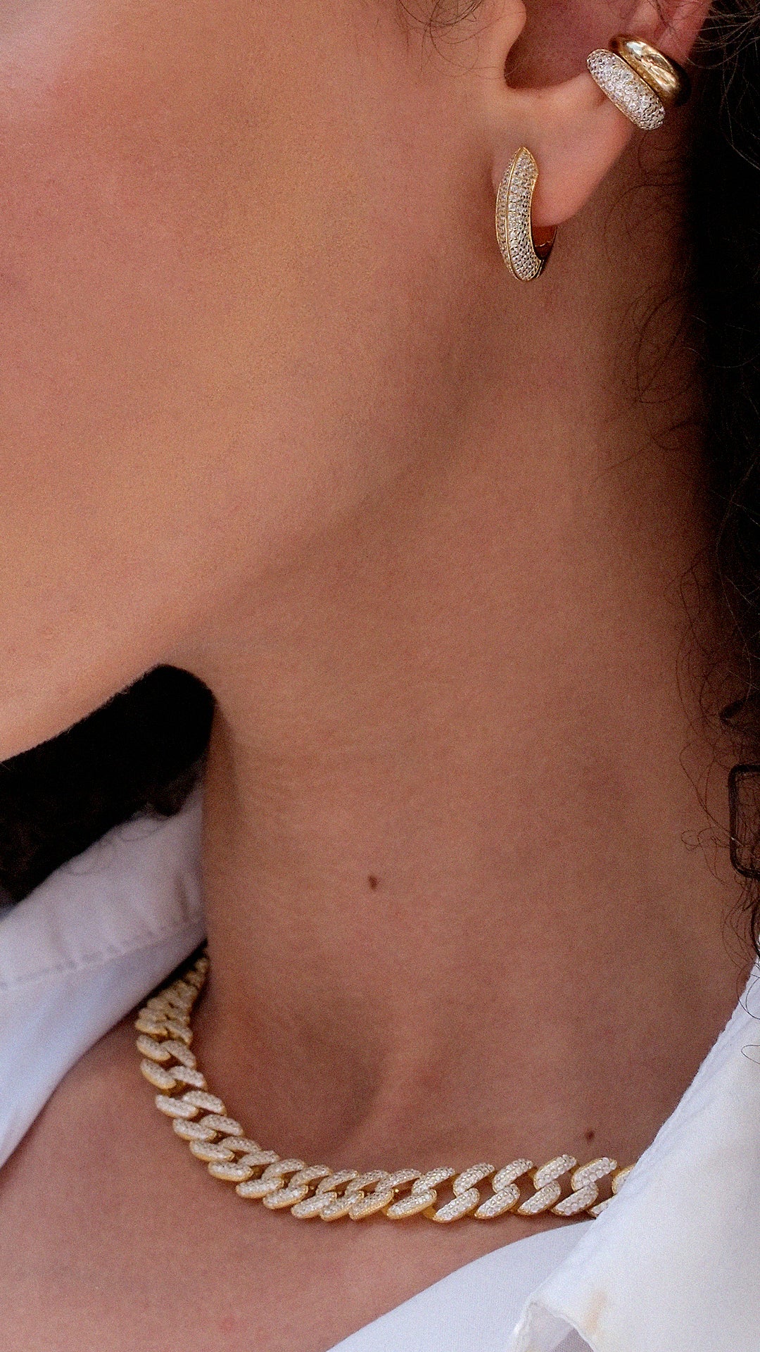 Close-up of stacked gold hoop earrings and a gold cuban chain necklace on a woman wearing a white shirt.
